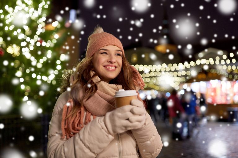 Joulu Lämpimästi pukeutunut nuori nainen hymyilee kuppi kädessä jouluvalaistulla kadulla.